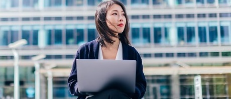 A young businesswoman sits in front of an office complex on a small wall with a laptop on her lap. She looks to the right as if she was looking into the future as a freelancer.