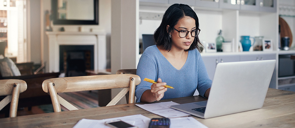 A woman sits in front of her laptop in her home office; she is very focussed on her work. Next to her, pens and papers lie on a table made of chic solid wood.